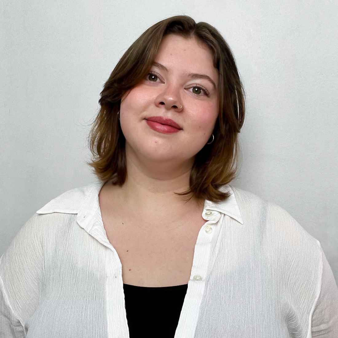 A young woman with a warm smile, brown hair parted in the middle, and red lipstick, wearing a white collared shirt over a black top against a plain light background.