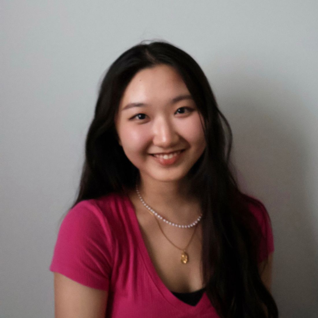 A smiling young woman with long dark hair wearing a pink top and layered necklaces, posing against a plain light background.