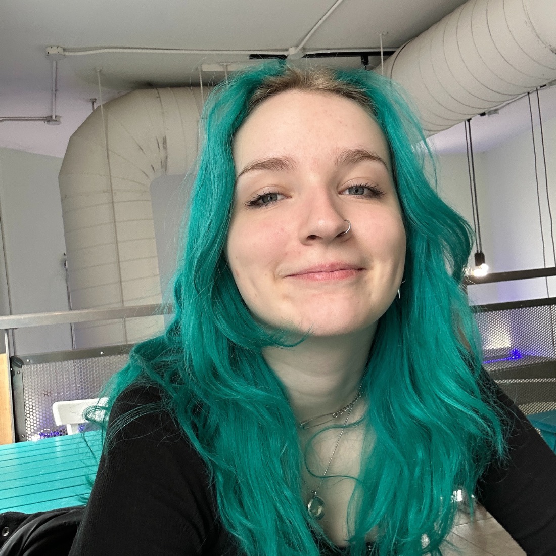 Young person with bright teal hair and a nose ring smiles warmly in an industrial setting with exposed pipes and lights. 