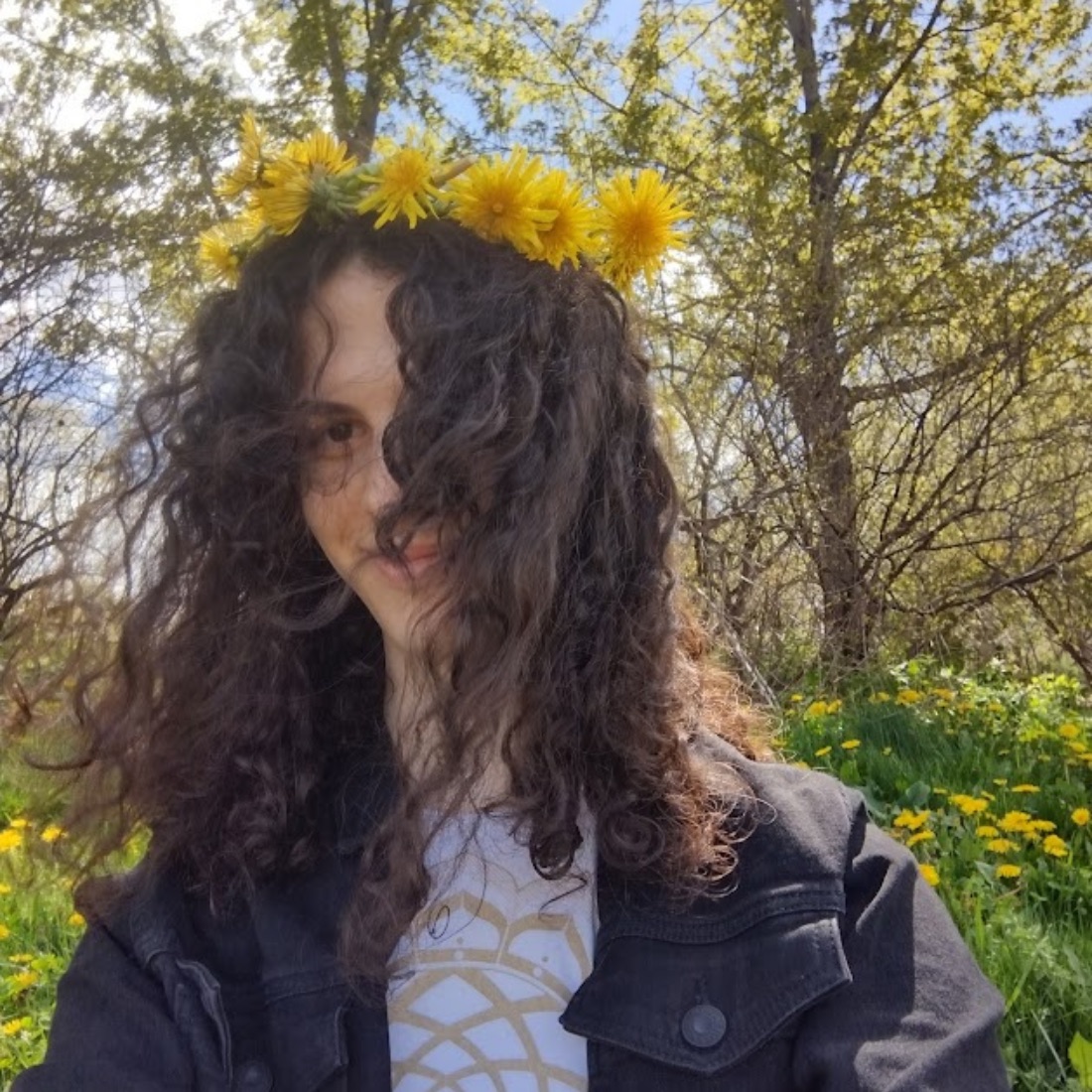 A person with curly hair, wearing a yellow flower crown, smiles subtly under the sunlight. They are in a field with blooming dandelions and trees.