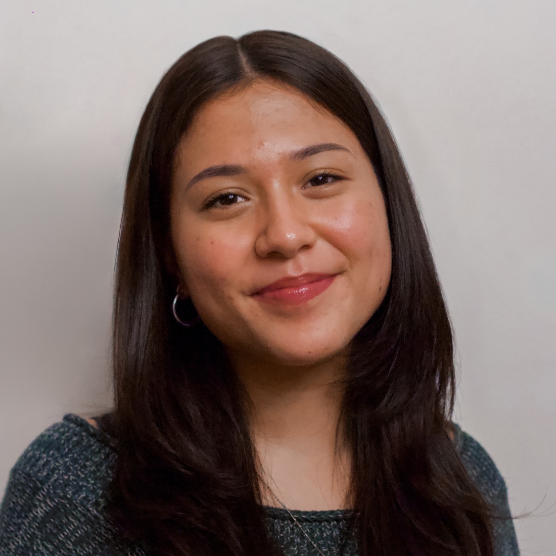 A young woman with long dark hair and a gentle smile is pictured against a plain background. She is wearing a dark sweater and hoop earrings.