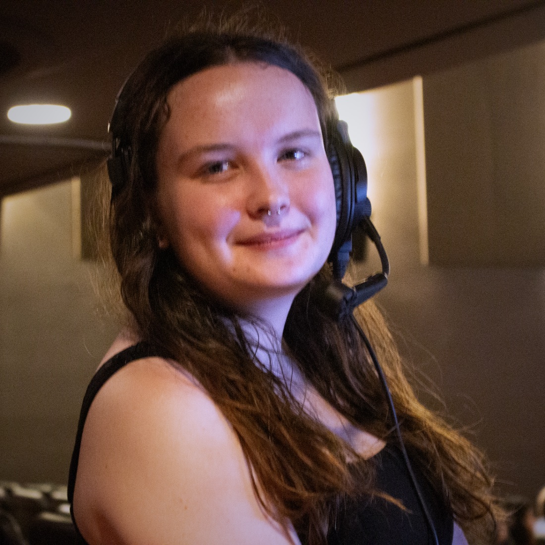 A person smiling, with long wavy brown hair, wearing a black headset in a dimly lit room with a relaxed atmosphere.