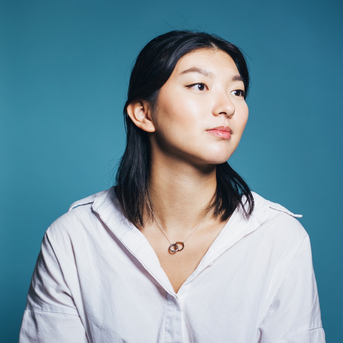 A woman with black shoulder-length hair, wearing a white shirt and a simple necklace. She looks away from the camera, to the right, set against a blue background.