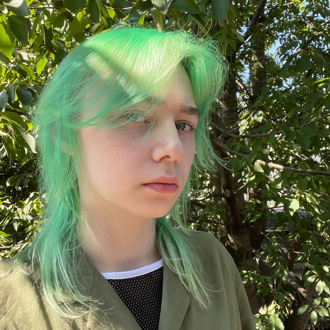 A person with bright green hair stands outside with a neutral expression. Sunlight filters through green leaves in the background, creating a natural ambiance.