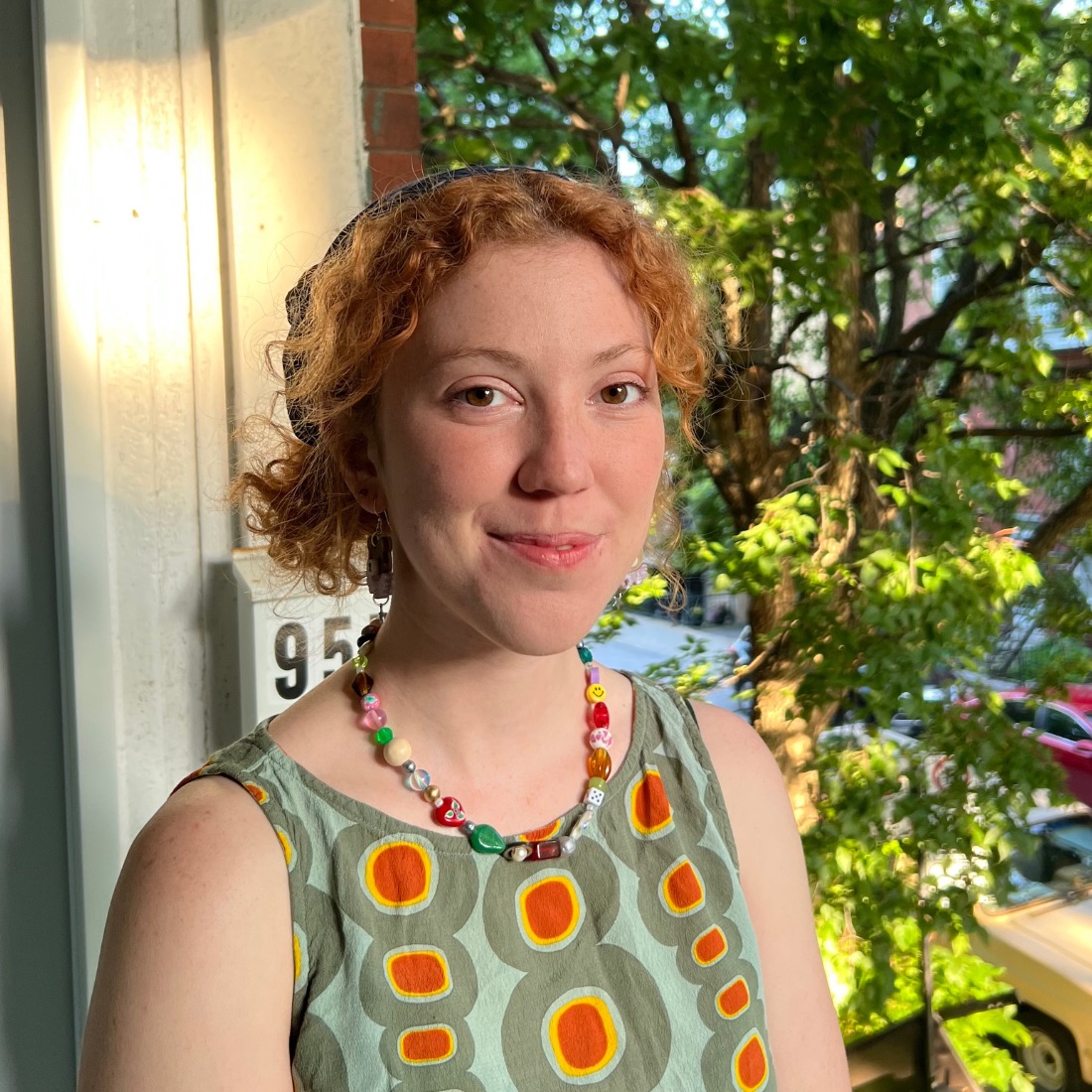 Young woman with short and curly red hair smiles on a sunlit porch. She wears a colorful dress and a beaded necklace. Lush trees and a city street in the background.