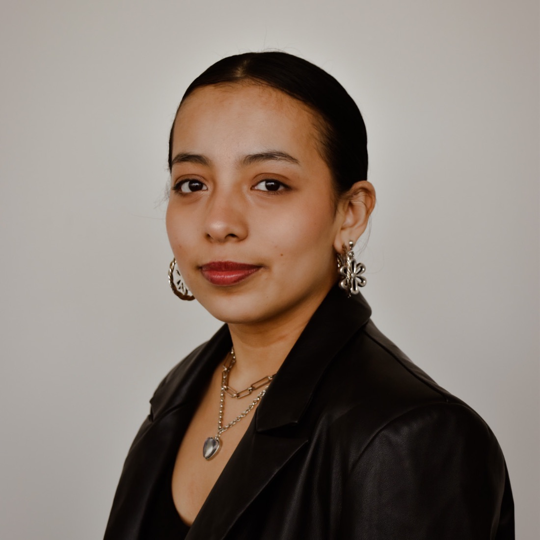 Young woman with hair pulled back in a bun, wearing a black leather jacket, hoop earrings, and layered necklaces. 