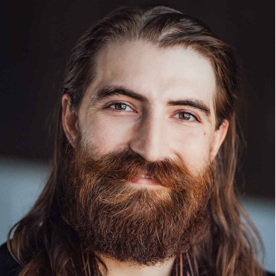 A close-up portrait of a smiling man with long dark hair and a thick, reddish-brown beard, set against a dark, softly lit background.
