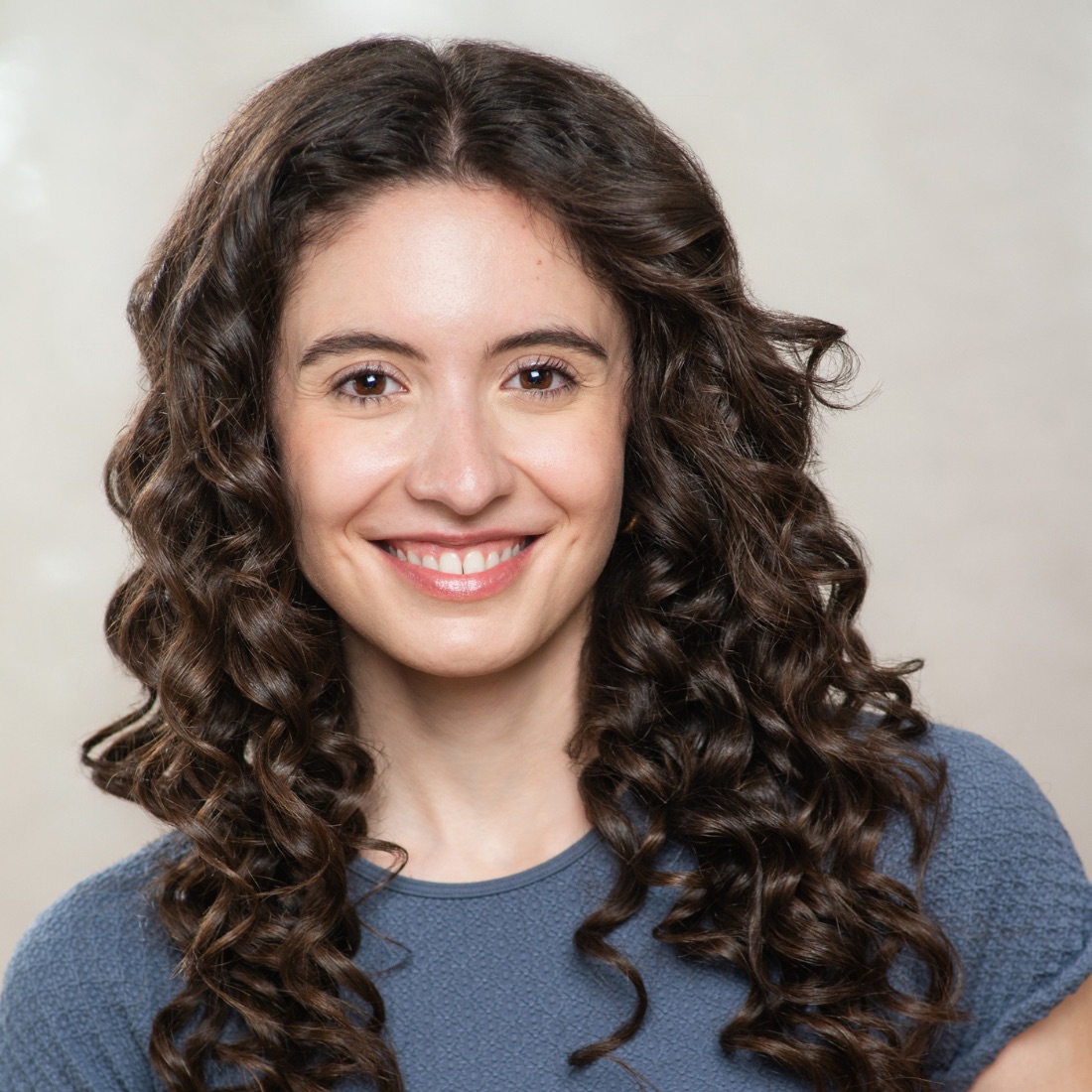 Smiling woman with long, curly brown hair wearing a blue top. She is facing the camera against a plain light background.