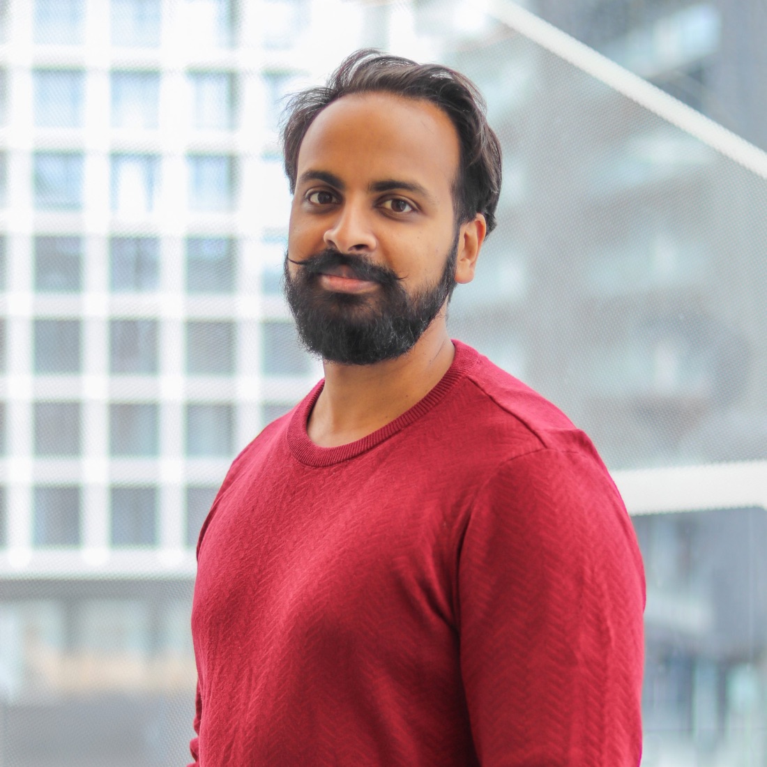 A man with a beard and mustache, wearing a red sweater, stands in front of a modern geometric window. 