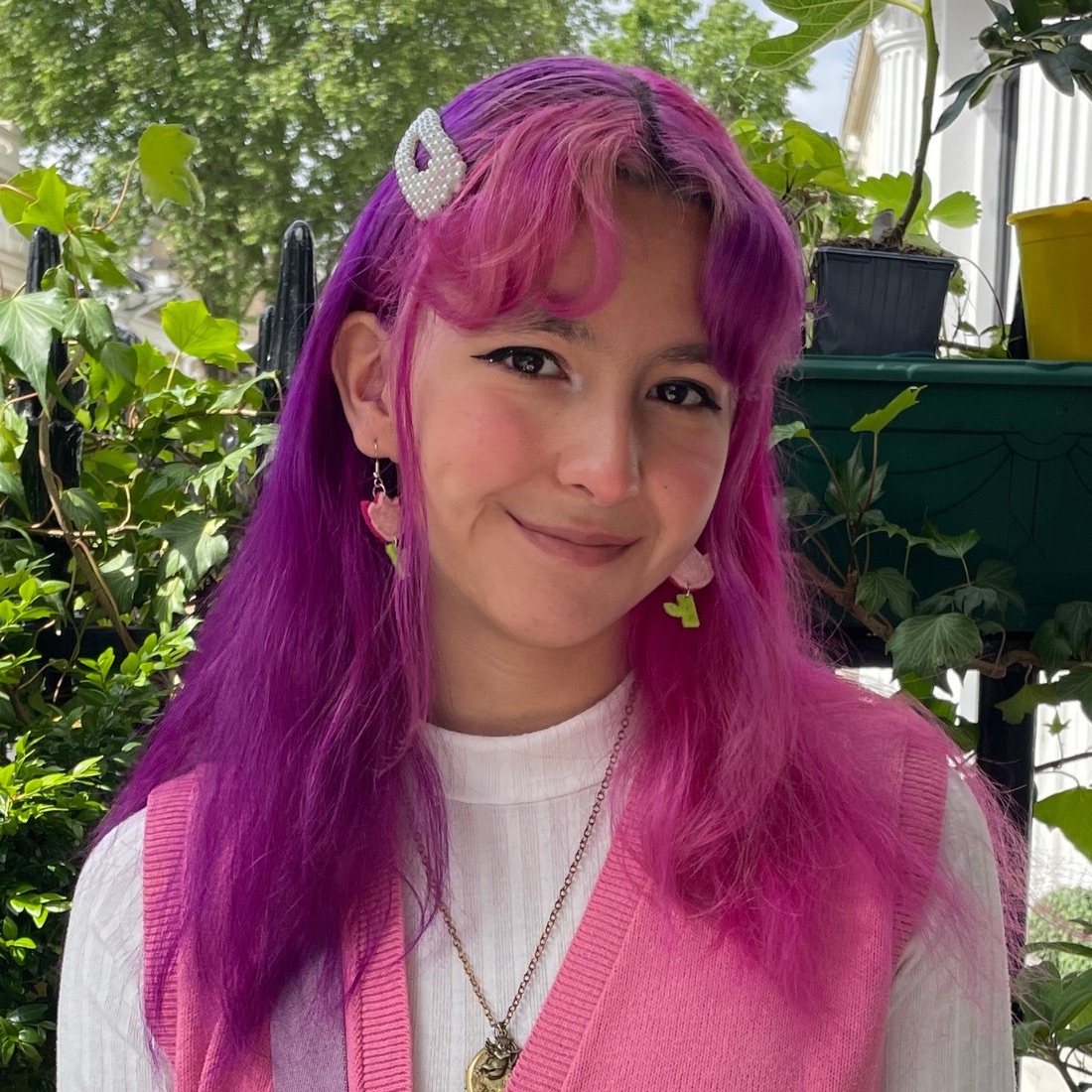 A young woman with vibrant pink-purple hair and a pearl hair clip smiles warmly at the camera. She wears pink earrings and a pink vest over a white top. Lush greenery in the background.