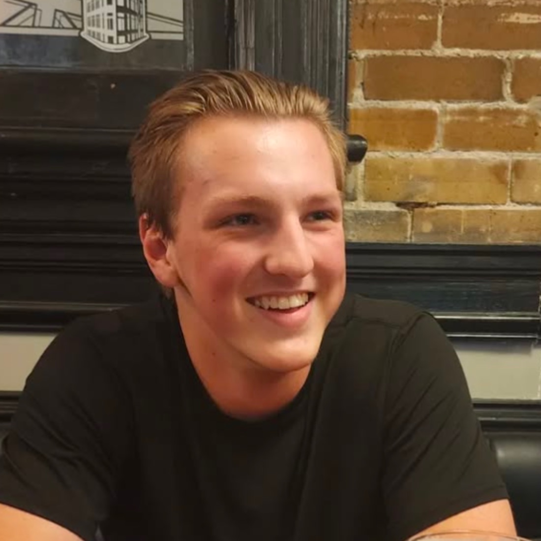 A young man with short hair, wearing a black shirt, is smiling warmly with his focus next to the camera. He sits against a brick wall.