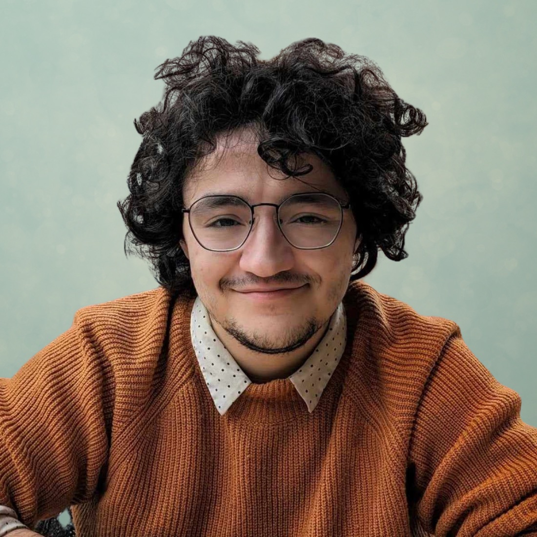 A smiling person with curly hair and glasses wearing a rust-colored sweater over a polka dot shirt in front of a soft green background.