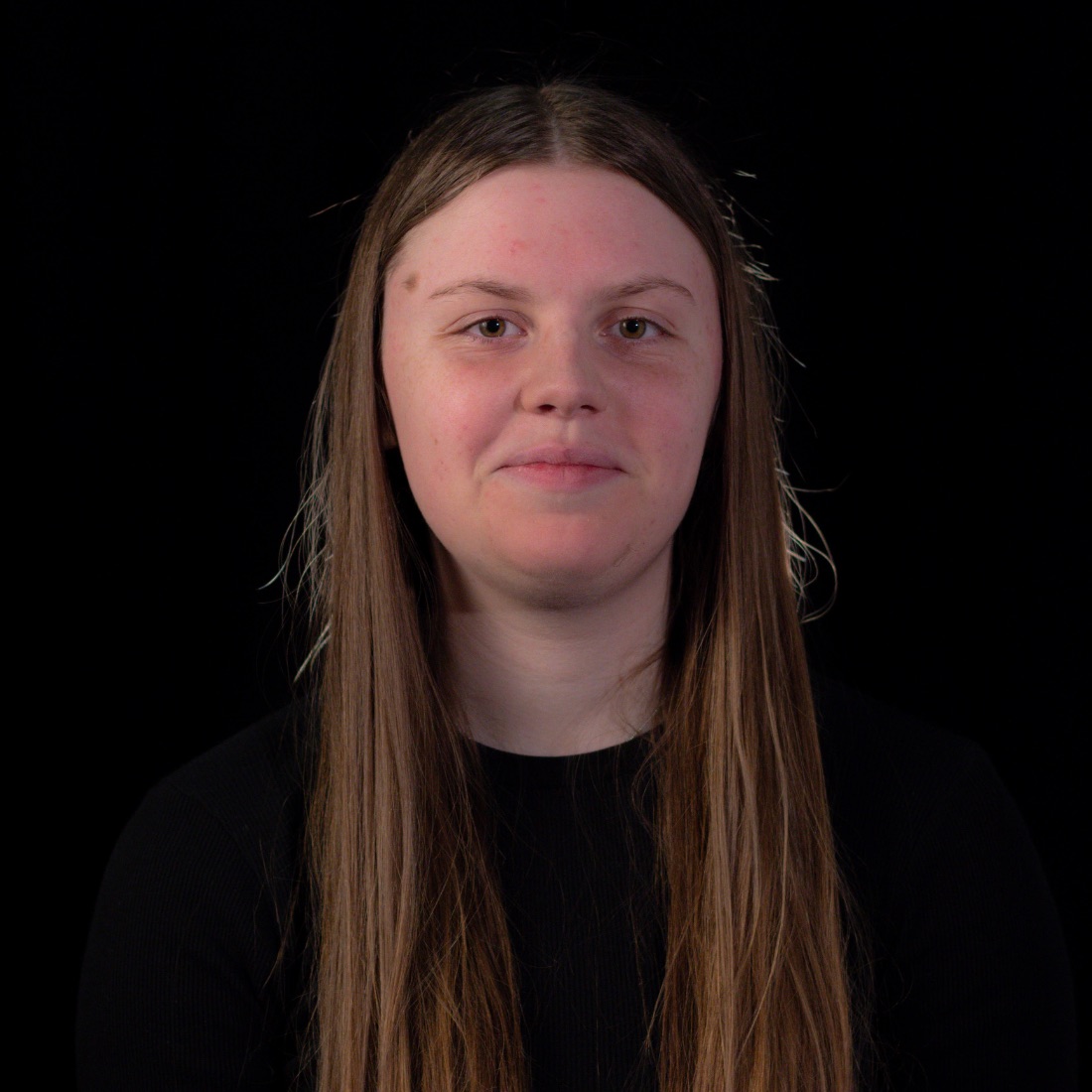 Young person with long light brown hair, wearing a black top, smiles subtly against a black background. 