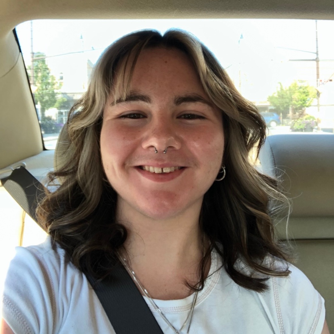  A person with wavy brown hair and a septum piercing smiles in the back seat of a car. 