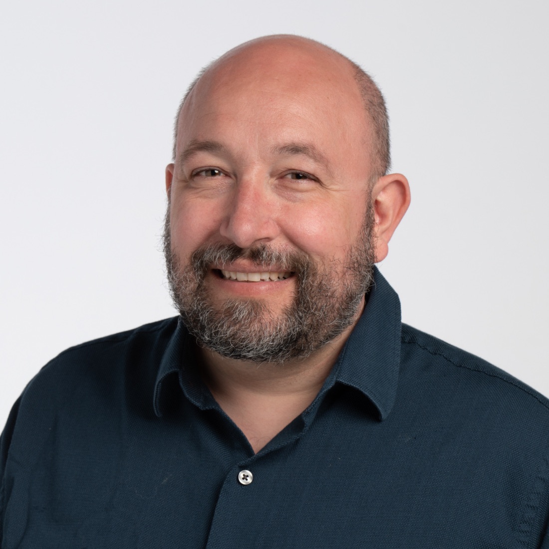 A bald man with a beard smiles warmly in a portrait against a plain white background, wearing a dark blue collared shirt