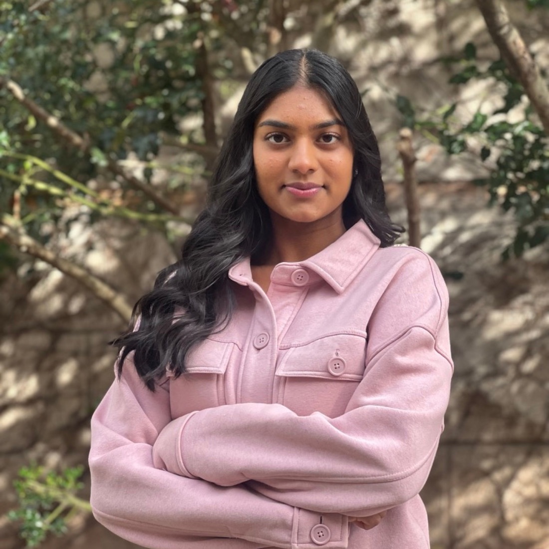 A woman in a pink jacket stands confidently with crossed arms in front of a blurred natural background.