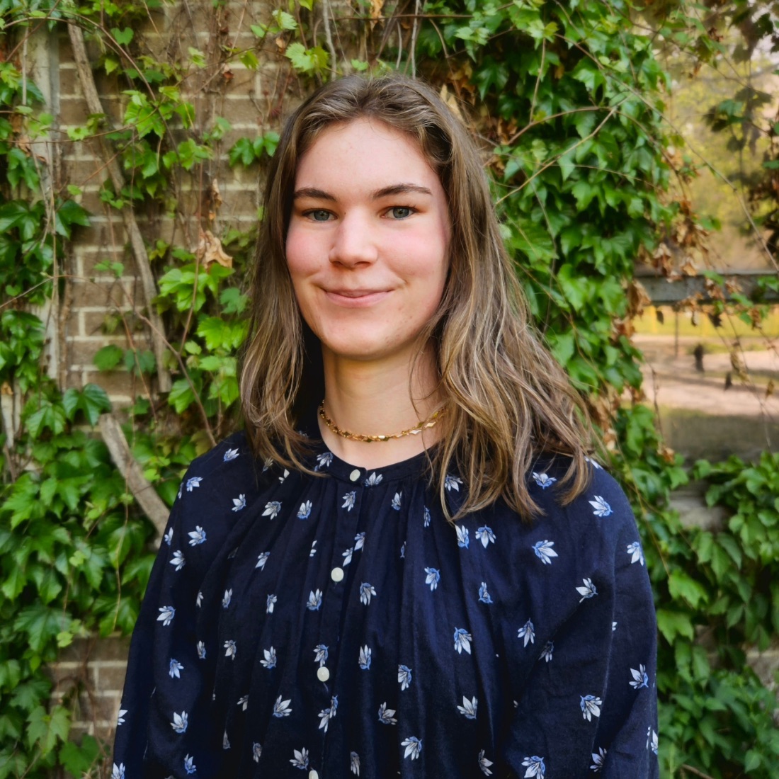 Young woman smiling softly in front of ivy-covered brick wall, wearing a dark floral blouse and a gold necklace. 