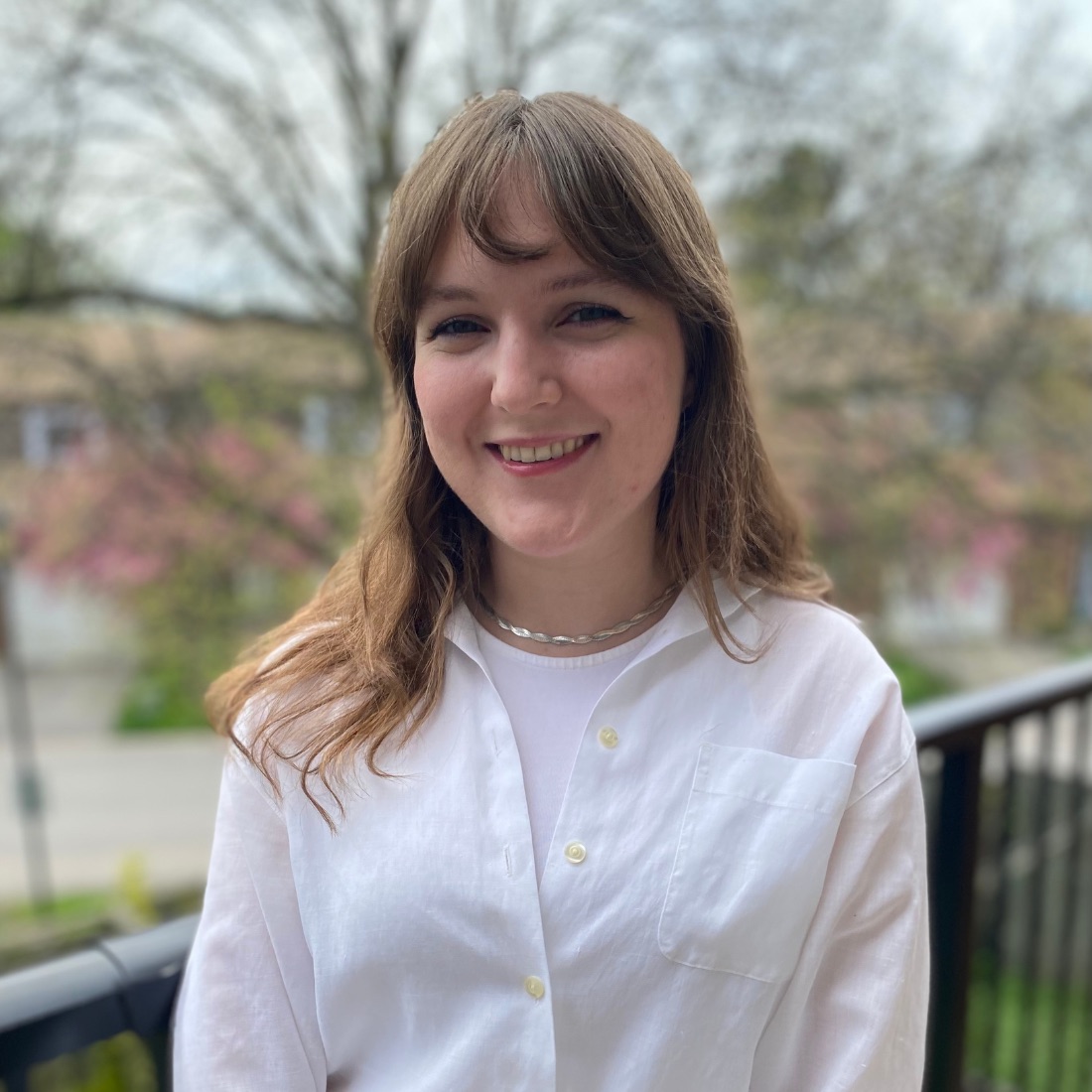Smiling woman with long brown hair and bangs, wearing a white button-up shirt, standing outside on a balcony with a blurry background of trees.
