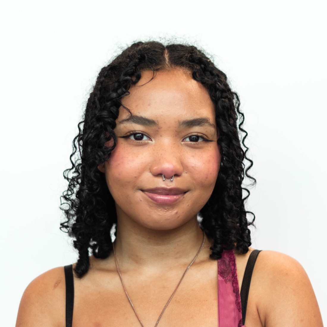 A woman with curly shoulder-length black hair and wearing a septum piercing and a pink top. The background is plain white.