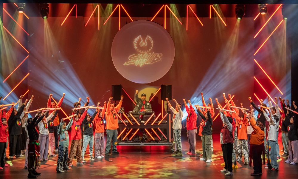 A wide shot of the stage, with colourful orange light bars and a projected logo illuminating the background. A large group of dancers stand on stage in a semi-circle with arms raised, surrounding a DJ at a console. Some dancers hold orange light tubes.
