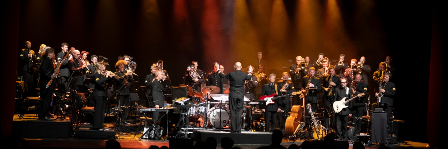 A large orchestra and band perform on stage under warm, colorful lights. The conductor stands center, facing musicians with brass, woodwinds, keyboards, and guitars.