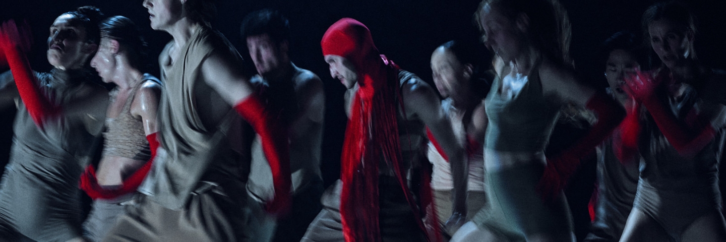 A dark, dynamic scene of a group of dancers in motion, clad in beige outfits and red gloves. The image is slightly blurry, with a focus on a dancer in a red headpiece in the center. 