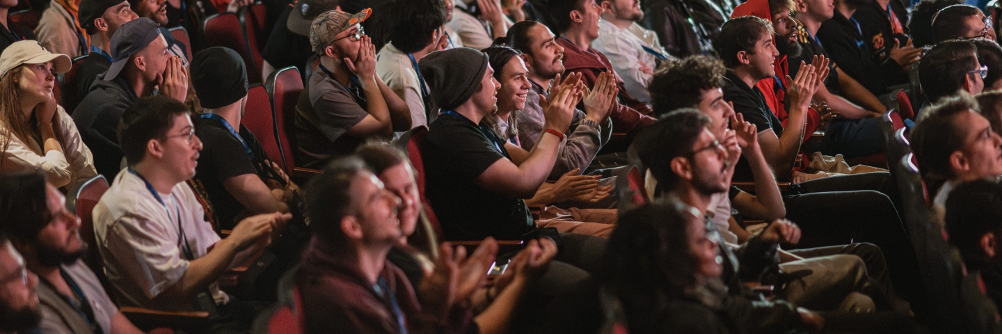 A diverse audience in rows of theatre seating is engaged and clapping, conveying excitement and appreciation at an event, creating a lively atmosphere.