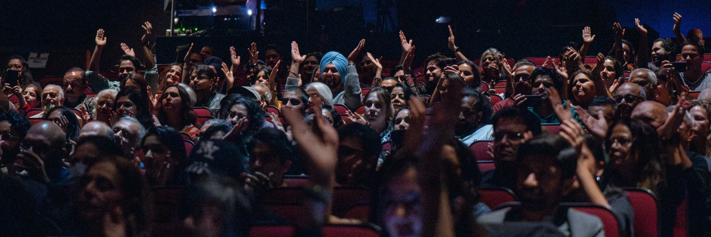 An audience in a dimly lit theatre, raising their hands in engagement and anticipation. Red seating is visible.
