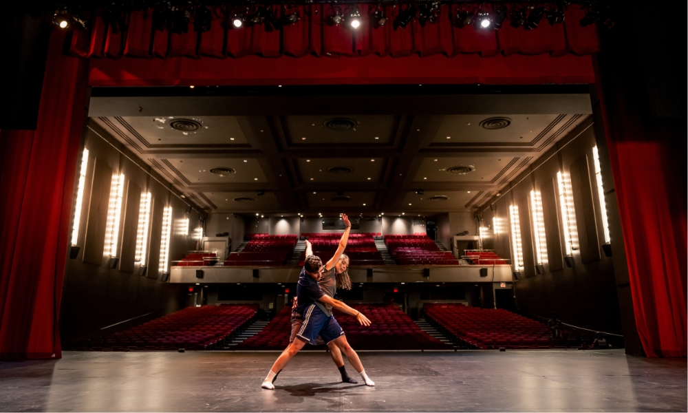 A female and male dancer duo perform on an empty theatre stage. They are facing away from the audience seating, and the shot features the empty theatre behind them. Red curtains frame the scene.