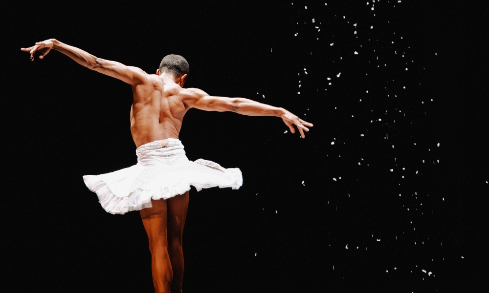 A male ballet dancer, wearing a white tutu and no shirt, is dancing on stage with arms extended. His back is to the camera, and he is surrounded by falling white snow against a black background.