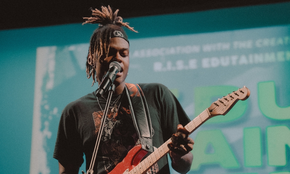 A musician with dreadlocks and wearing a black graphic t-shirt passionately plays an electric guitar and sings into a microphone on stage, set against a backdrop with vibrant blue graphics and text.