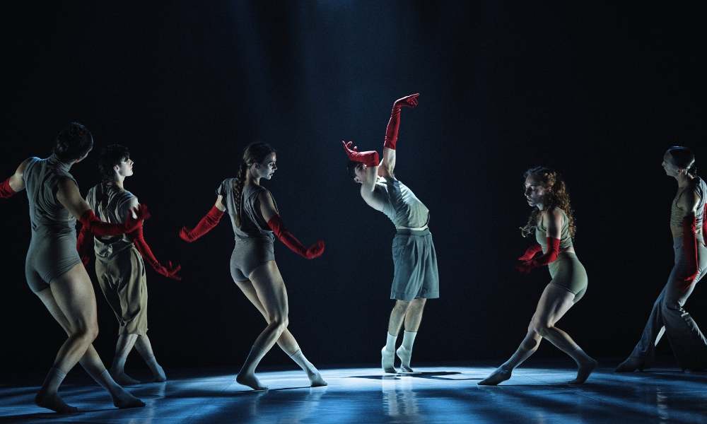 A group of six dancers perform on stage, dressed in neutral outfits with red gloves. They strike dynamic poses under a dramatic spotlight, conveying intensity.