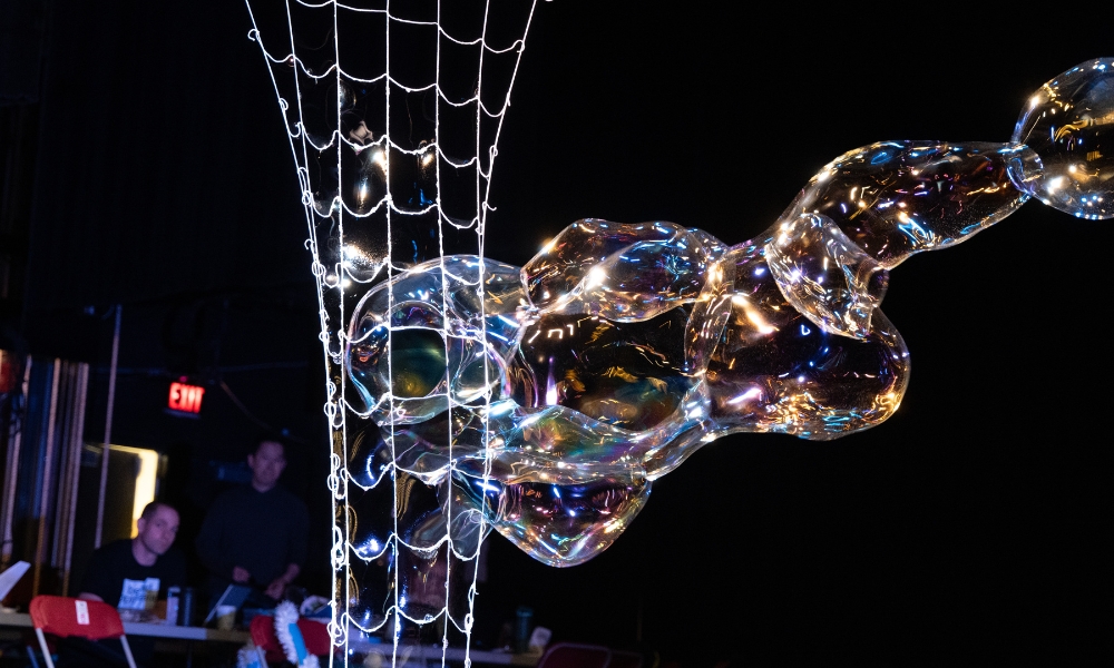  A cluster of iridescent bubbles is caught in a string net, illuminated by colorful lights, against a dark background. Two people observe from the background.