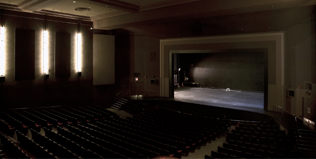 A wide shot of an empty theatre with rows of red seats facing the stage and proscenium arch. Soft lighting on side walls creates a quiet, anticipatory atmosphere.
