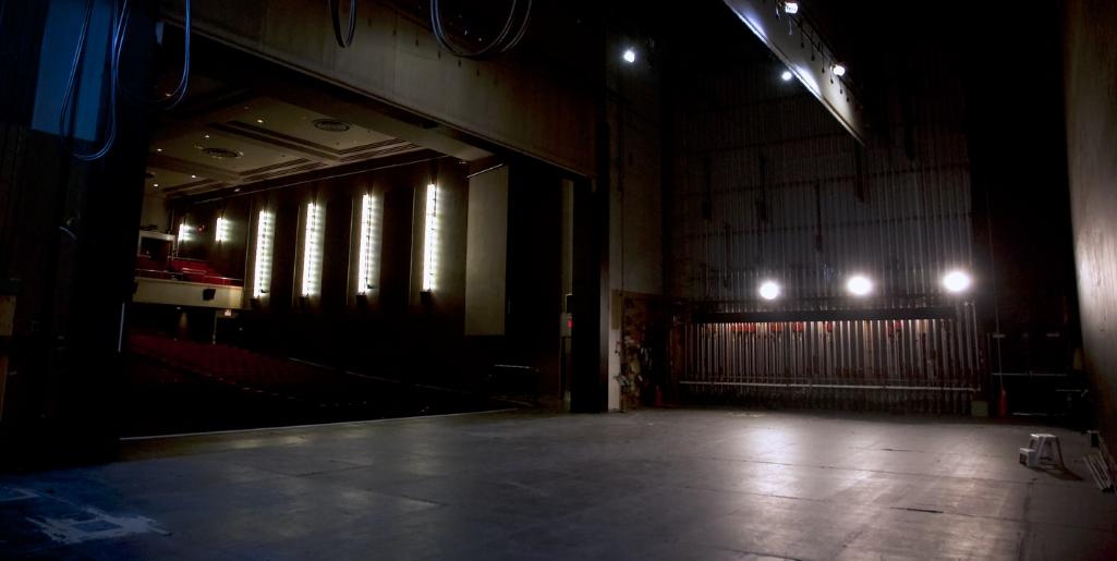 A backstage view of the stage looking towards an empty theatre. The stage is dark and empty, showing the large and spacious floor. In the back, the theatre’s fly rail is featured. 