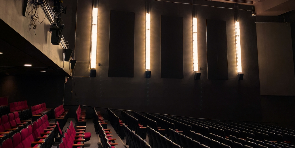 A side shot of a theatre with rows of empty red seats and vertical wall lights creating a warm ambiance. Black sound panels add to the sleek, modern look.