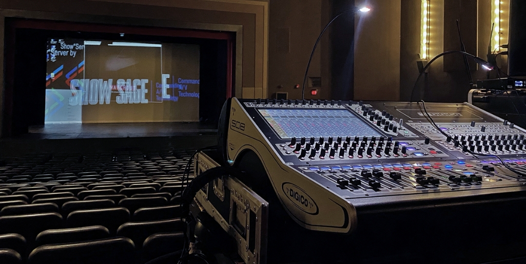 A sound console at the back of an empty theatre. The stage is in the background with a partially visible projection screen. Empty rows of seats fill the foreground. The photo is from the perspective of a sound technician.