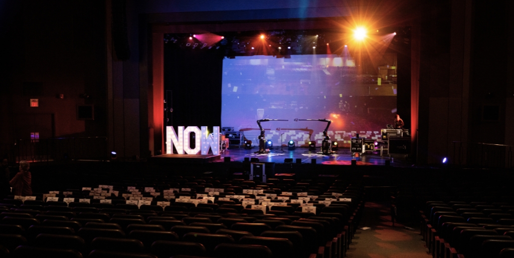  A wide shot of a theatre stage with colourful stage lights. On stage, there are large, illuminated letters spelling "NOW". Next to the letters, there are two black Kuka robot arms.