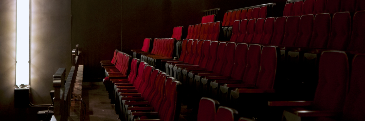 Dimly lit auditorium with rows of empty red seats, creating a quiet and expectant atmosphere. A soft glow from a vertical light on the left illuminates the wall.