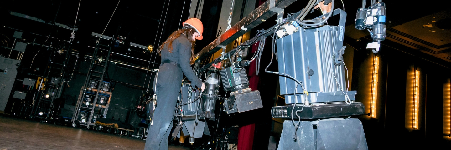 A stage crew member in an orange hard hat adjusts lighting fixtures on a dark theatre stage. The shot is from a low angle.