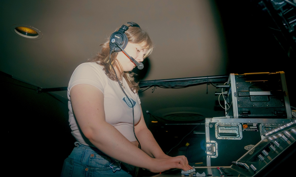 A sound technician wearing a headset operates a sound board under dim lighting. They focus intently, suggesting concentration and professionalism in a technical setting.
