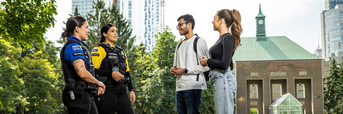 Two TMU Security guards speak with two students in the Quad.