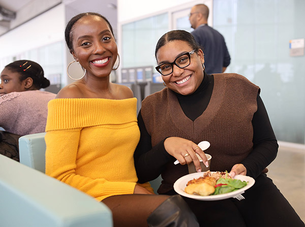 Black staff, Aneka Olbino and Bianca Marryshow, eating food and smiling at the Black Excellence Mixer. 