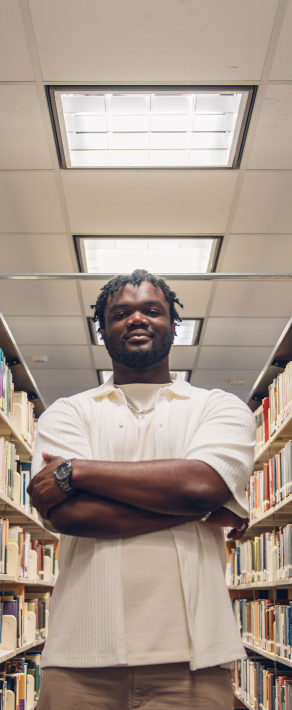 Black student, Divine Amayo, smiling in the middle of an aisle of books in TMU’s library. 