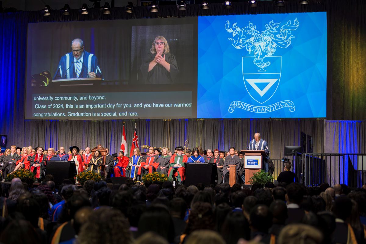 A large screen is positioned directly above the Convocation stage. It is showing a live feed of the stage, a live ASL interpreter, live captions of the ceremony and an image of the TMU shield. 