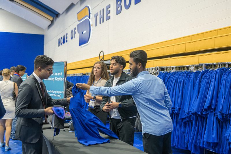 3 convocation staff members are handing a blue bachelors gown to a graduand