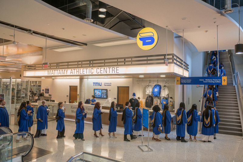 A single file line of grads in blue gowns walking up a set of stairs to the 3rd floor of the Mattamy Athletic Centre