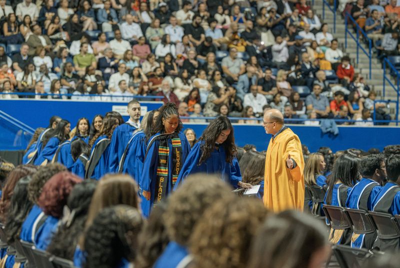 A volunteer in a gold gown directs a line of grads to their seats in the arena