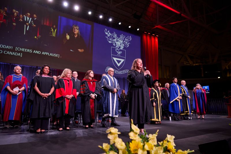 A person wearing a black gown is standing in the centre of the stage with a mic and singing O'Canada