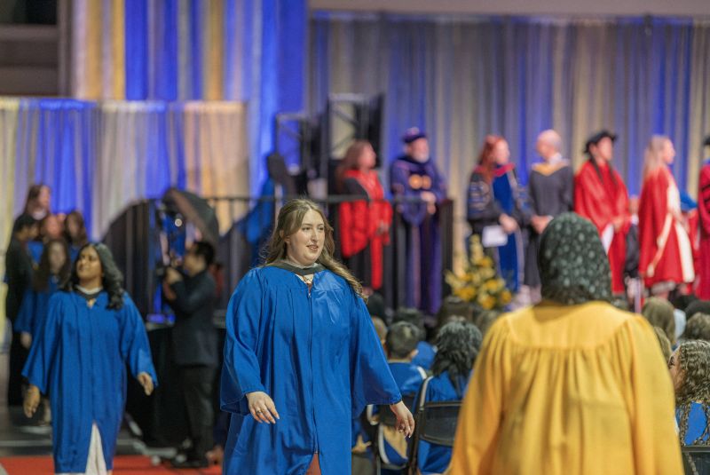 Grads in blue gowns are directed back to their seats after crossing the stage by a staff member in a gold gown